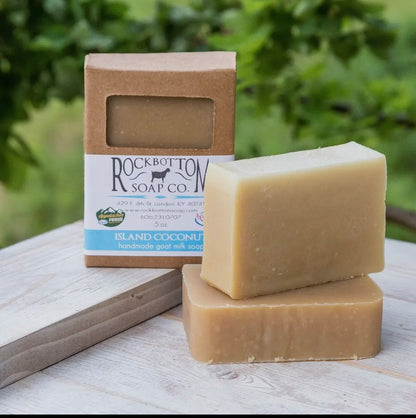 Two bars of Island Coconut soap from Rockbottom Soap Co. on a wooden surface with a blurred green background.